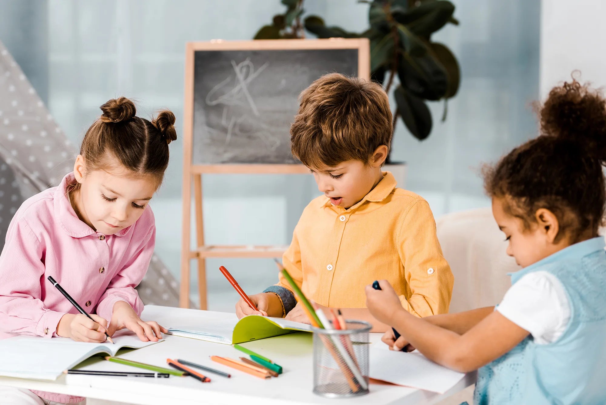 Three children sitting at a table with books and colored pencils, with a chalkboard in the background.
